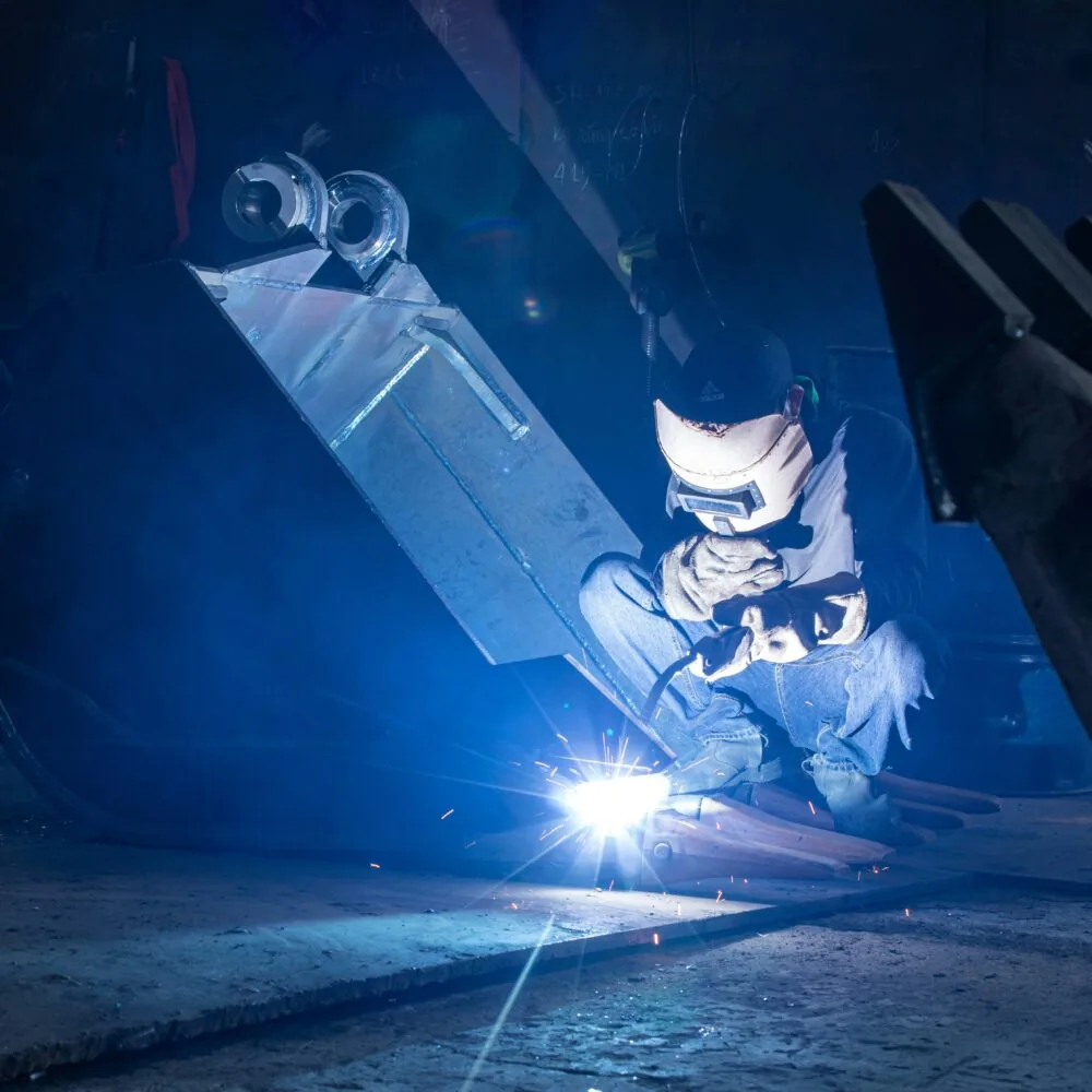 Ein Schweißer mit Schutzhelm arbeitet an einer Metallkonstruktion, umgeben vom blauen Licht des Lichtbogens.